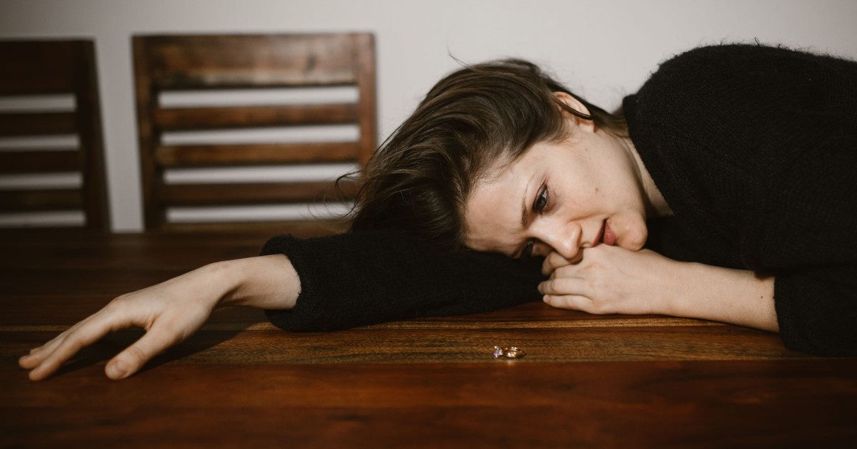 A woman lying on a wooden table, reflecting on a wedding ring, portraying emotions of sadness and contemplation.