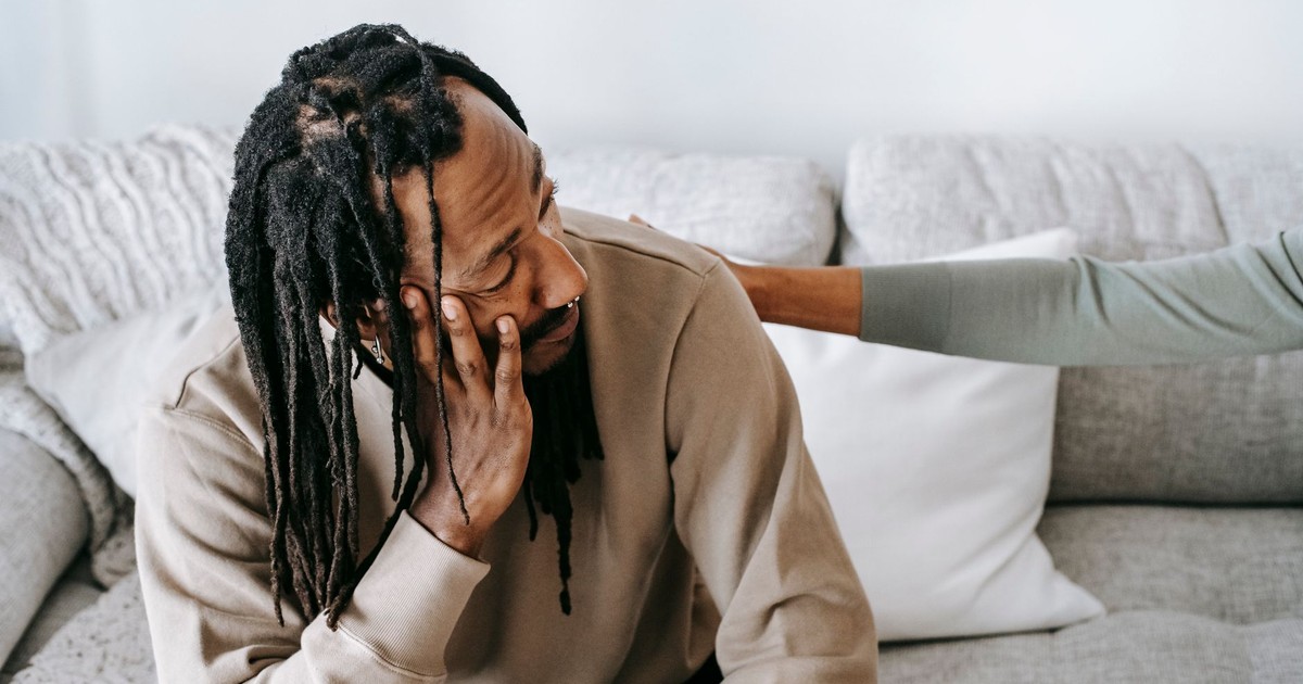 Depressed African American male in uneasy situation discussing problem with female psychologist or friend while sitting on sofa together