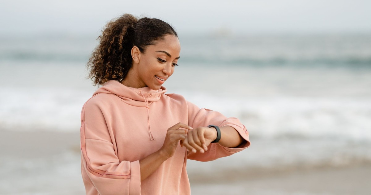 Smiling woman in a pink hoodie checks her smartwatch by the ocean.