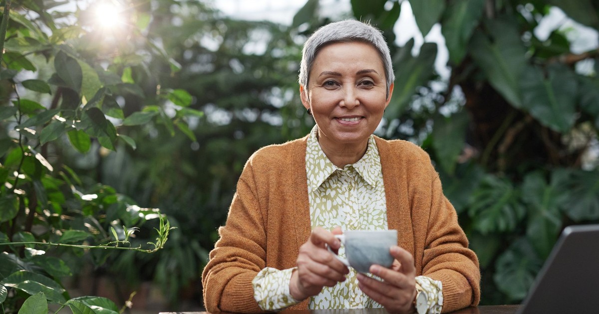 Senior woman with a coffee cup smiling in a lush greenhouse while working on a laptop.