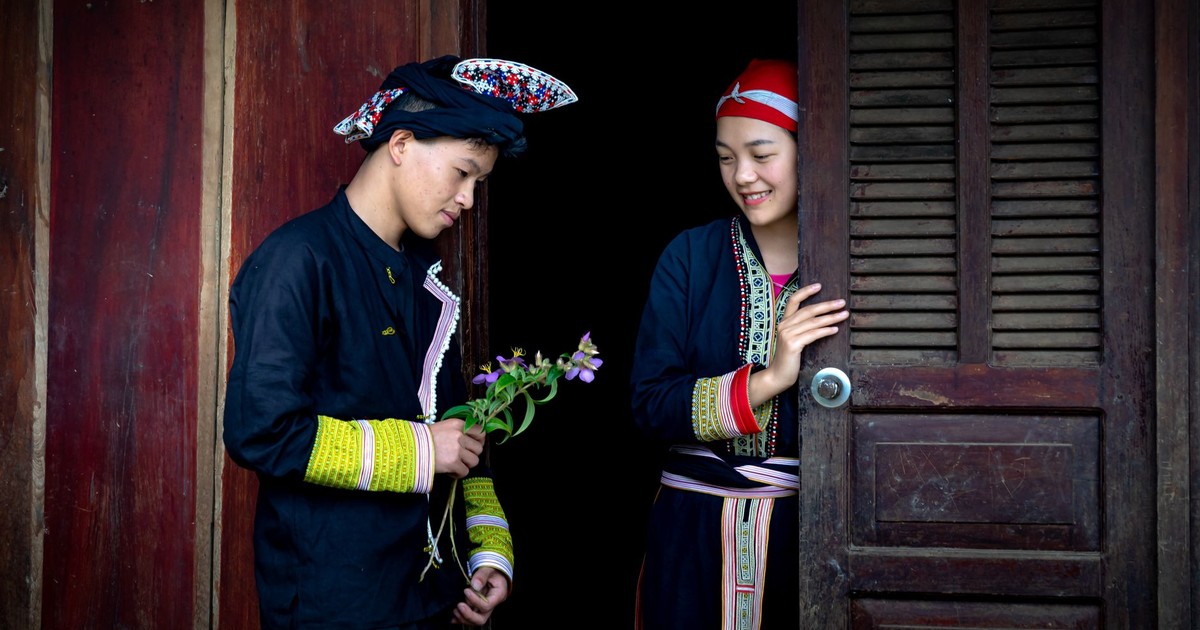 A man presents flowers to a smiling woman at a wooden doorway, highlighting cultural attire.
