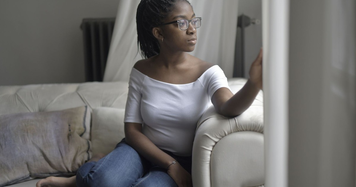 Woman in glasses relaxing on a sofa near window, deep in thought and looking outside.