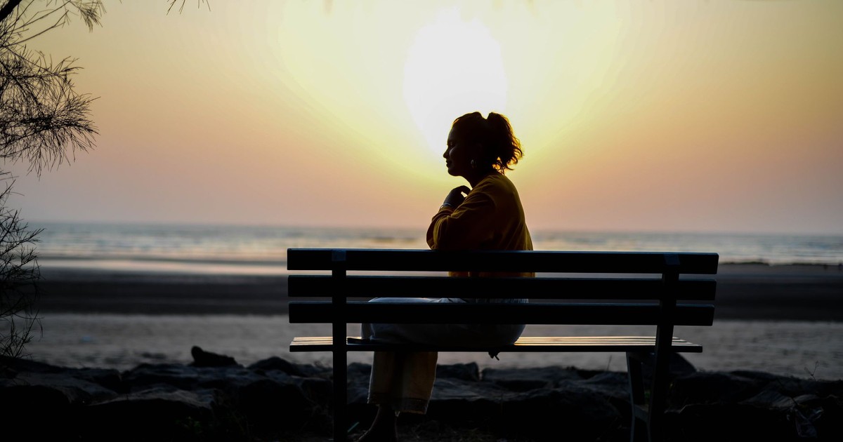 A silhouette of a woman sitting on a bench by the beach during sunrise, capturing solitude and contemplation.