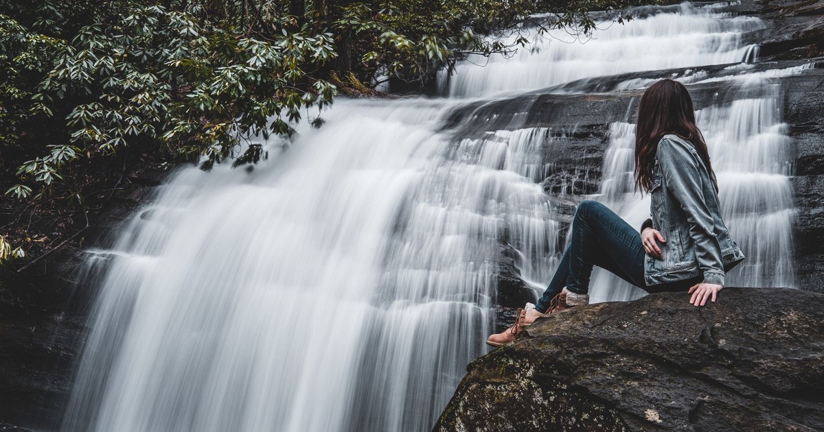 A woman in a jacket sits on a rock by a flowing waterfall surrounded by trees.