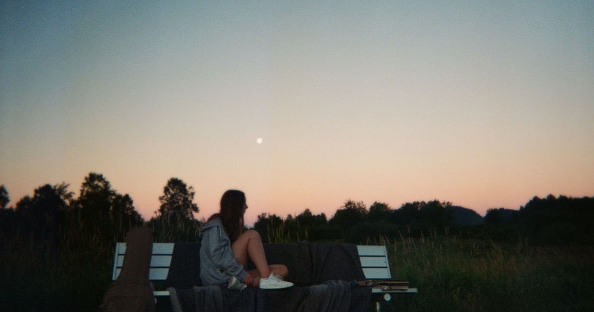 A woman sits on a bench at twilight, surrounded by nature with a guitar nearby.
