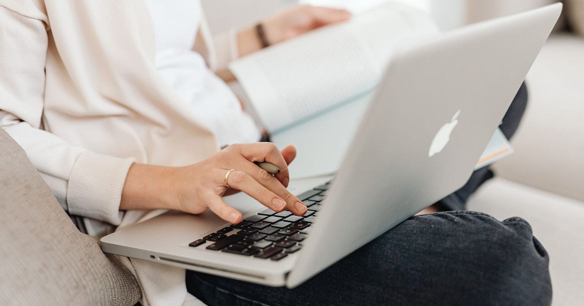 Close-up of a person multitasking, typing on a laptop while holding a book on their lap.
