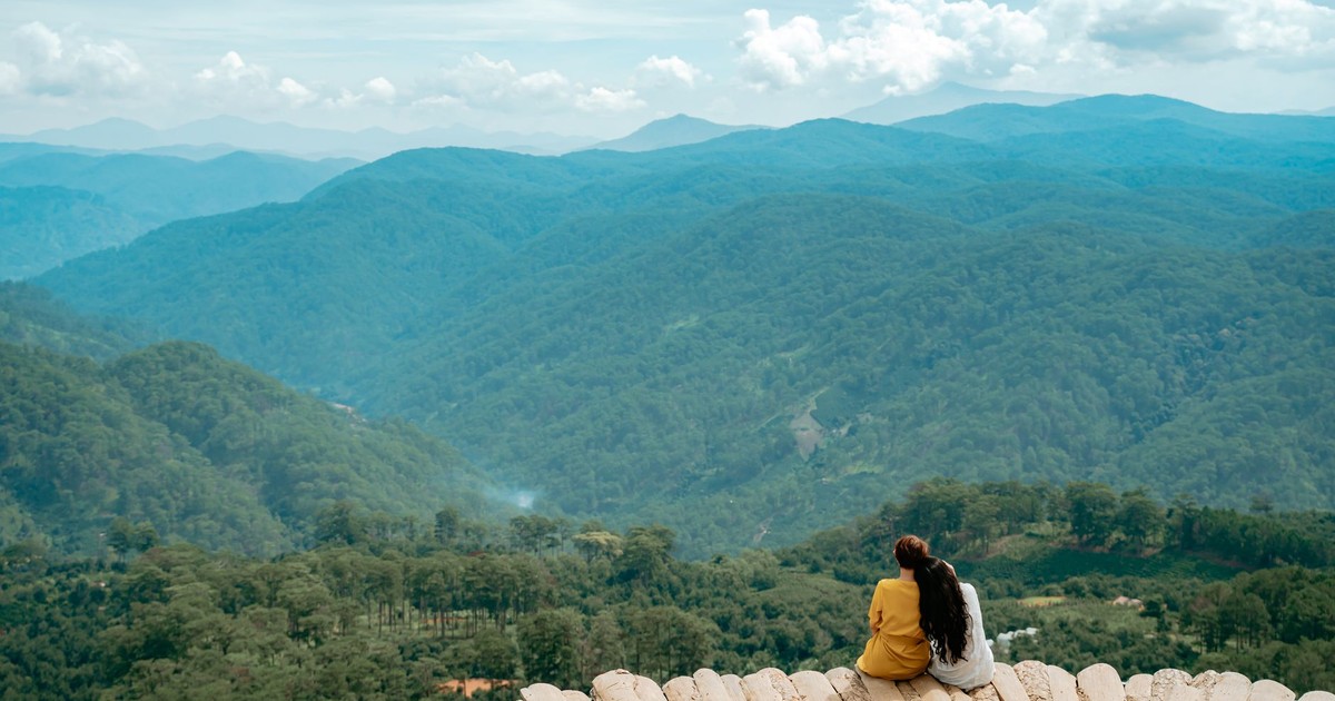Back view of anonymous tourists cuddling while sitting on viewpoint and admiring picturesque mountain valley covered with lush green vegetation