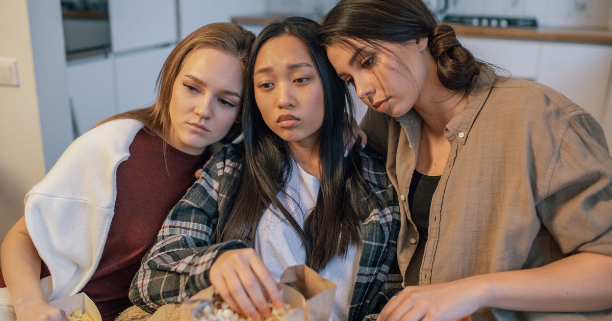 Three young women consoling each other while sharing popcorn indoors, signifying friendship and support.