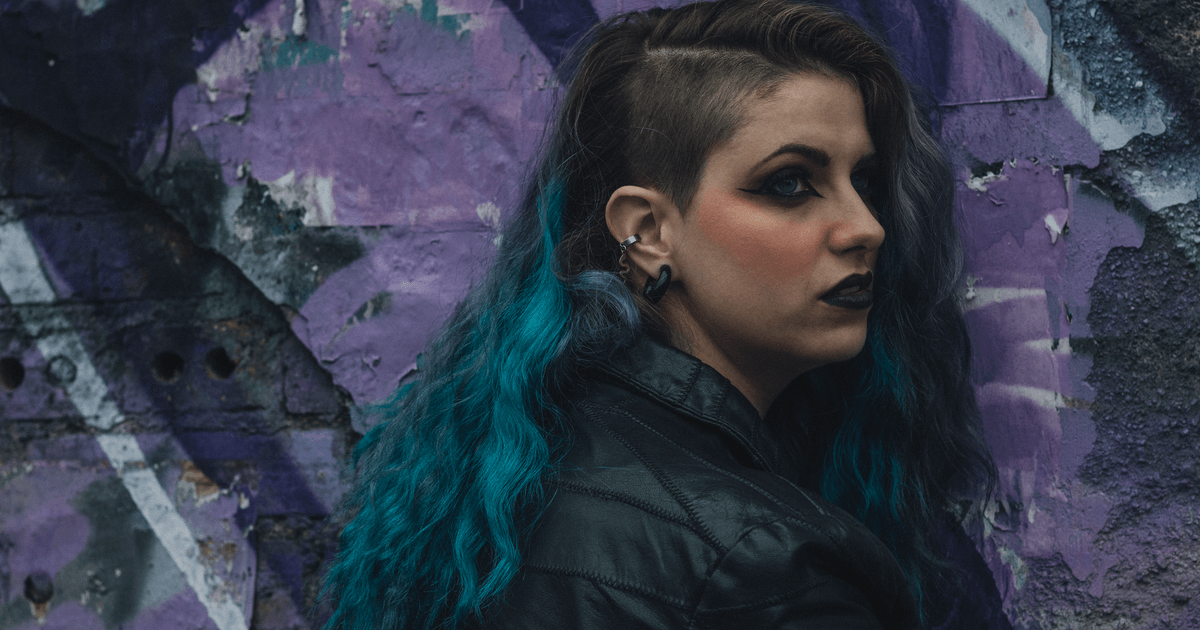 Portrait of a woman with blue hair against a graffiti wall, showcasing urban and alternative fashion.