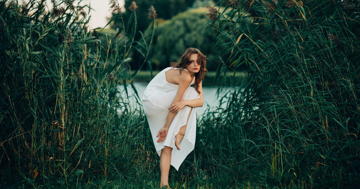 Woman in a white dress posing gracefully by lakeside reeds.