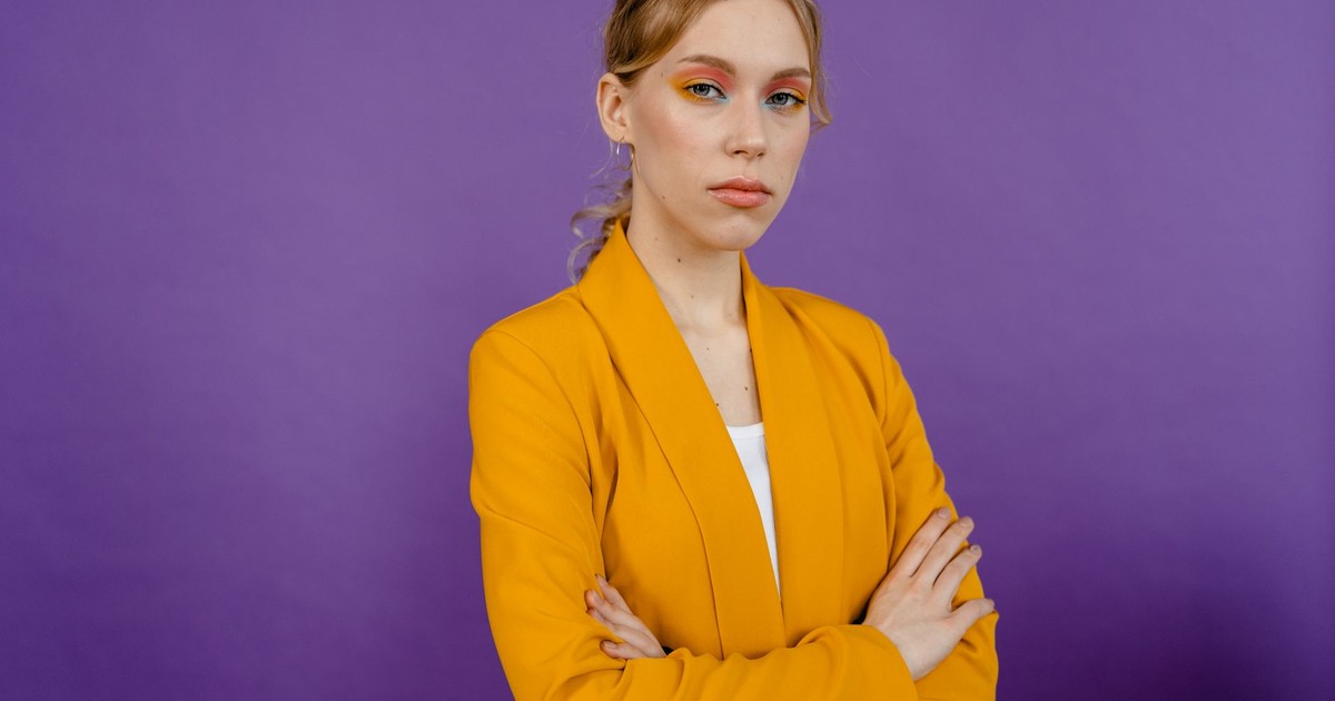 Portrait of a confident woman in a yellow blazer with colorful eye makeup against a purple background.