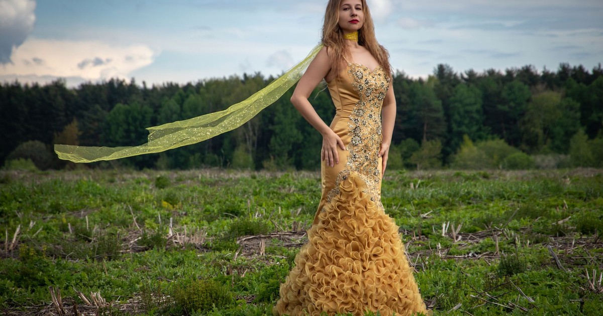 A woman in a golden evening gown stands confidently in an open field under a cloudy sky.