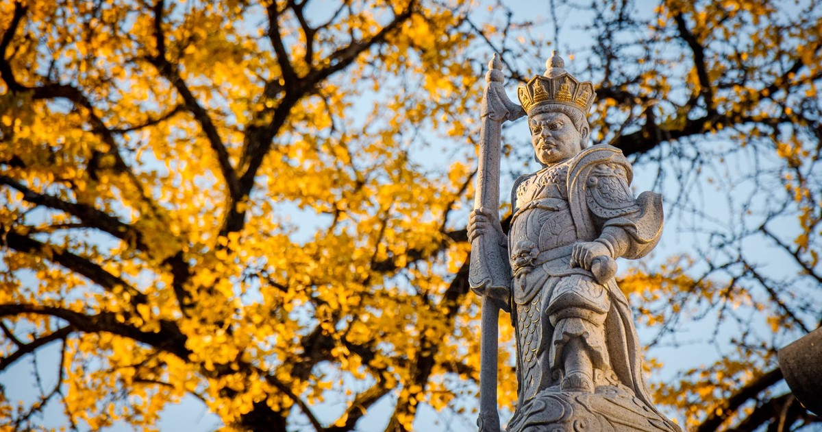 A detailed stone statue of a warrior in a crown stands against golden autumn leaves.