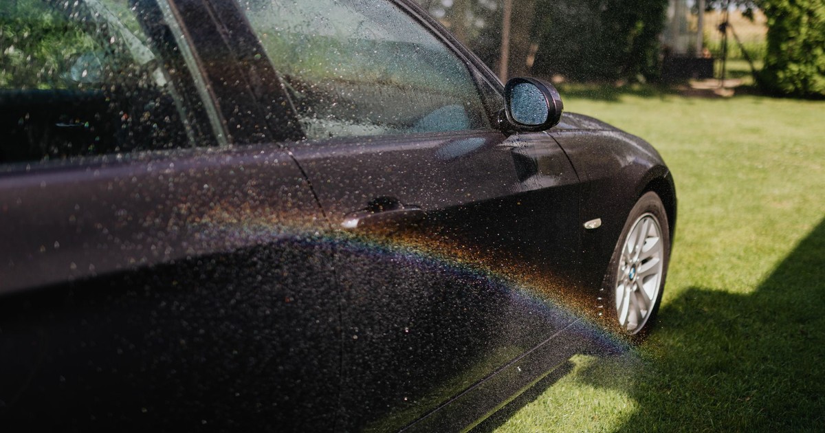 A black car being washed, capturing a rainbow in sunlight with water droplets on the exterior.