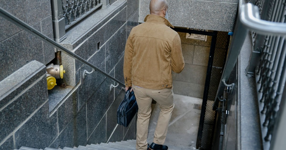 A bald man in a tan coat descends stairs carrying a briefcase in an urban setting.