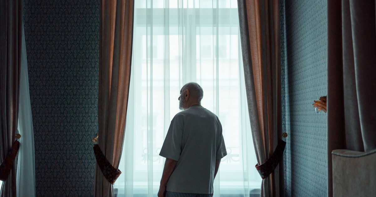 Elderly man standing by a window in an elegant room, looking outside. Ideal for concepts of isolation or contemplation.