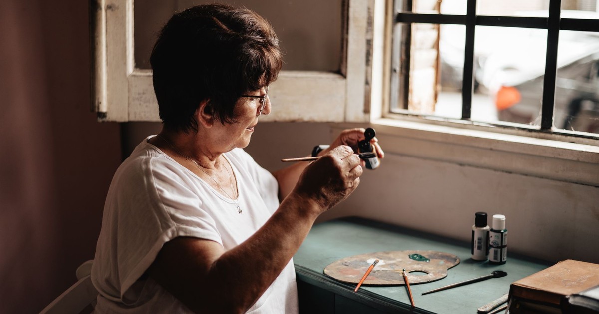 Senior woman engaged in painting by a window, with art supplies on the desk, creating art in natural light.