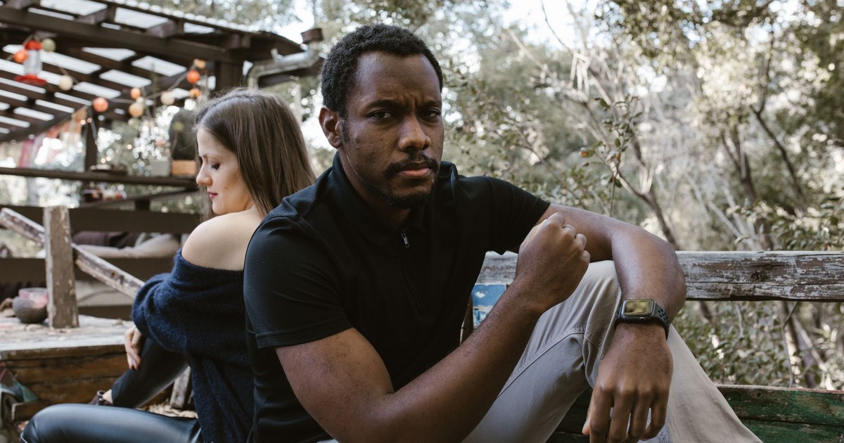 A thoughtful man sits outdoors, with a woman sitting distantly behind him.