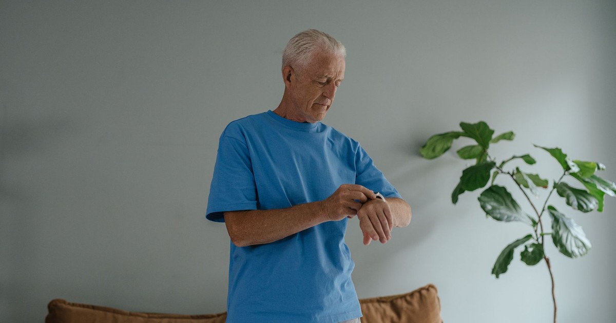 Elderly man in a blue shirt checking his wristwatch indoors by a plant.