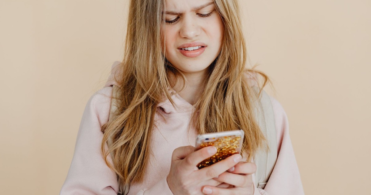 Teen girl with blonde hair in pink hoodie using smartphone, expressing curiosity indoors.