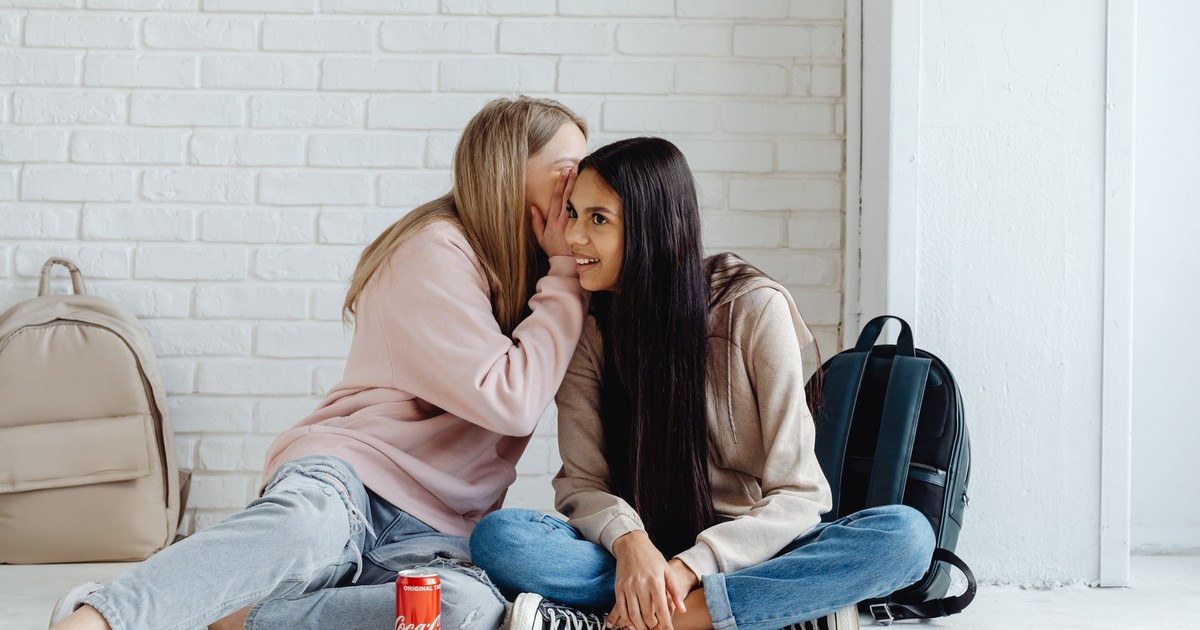 Two teenage girls whispering and sharing secrets while sitting indoors, surrounded by backpacks.