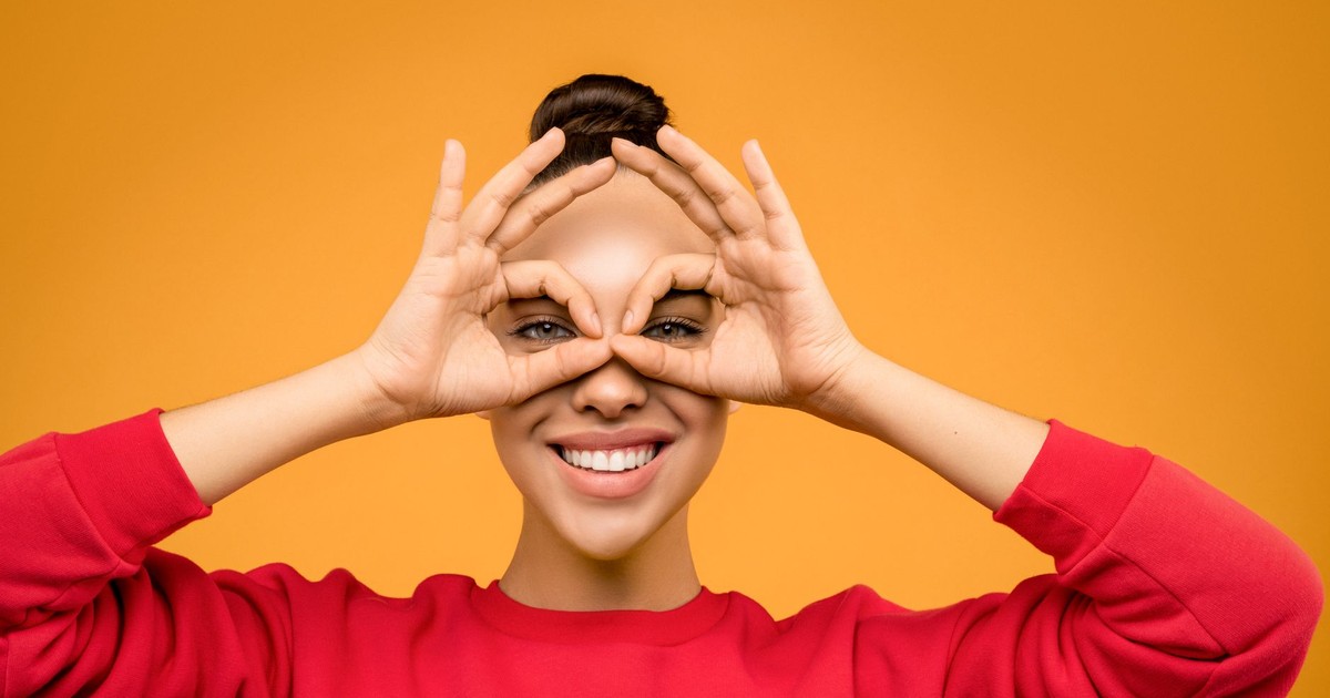 Cheerful woman making binoculars with hands, smiling against orange backdrop.
