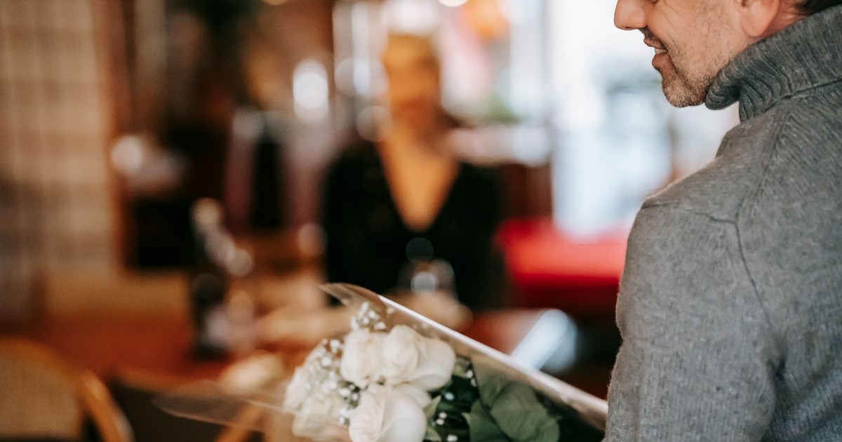 Middle aged couple having dinner in light room while male standing with bouquet with roses near table and chairs