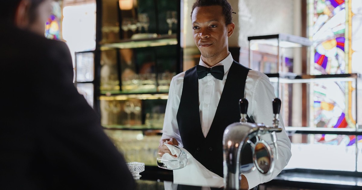 A bartender in formal attire serves a customer in a classic, stylish bar setting.
