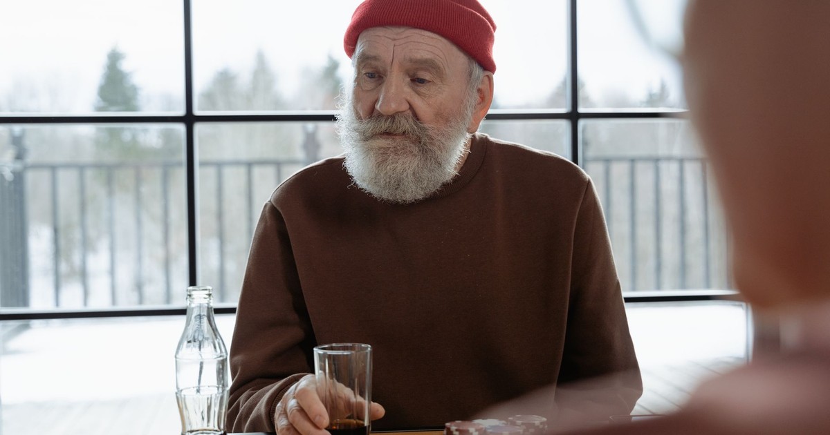 Senior man playing cards at a table indoors with a drink, relaxed moment.