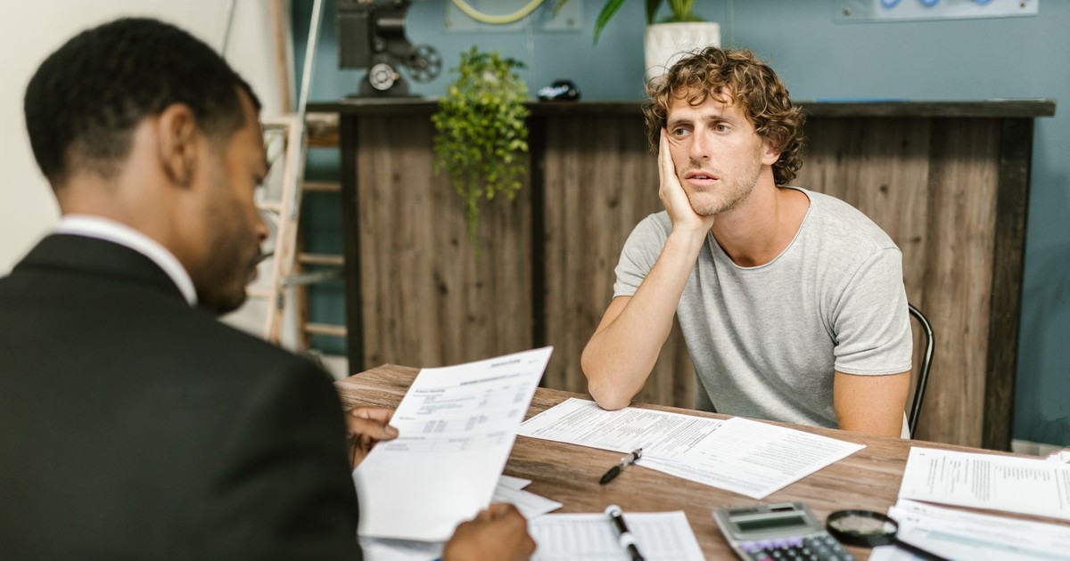 Man in office, appearing stressed during paperwork discussion, sitting opposite professional.