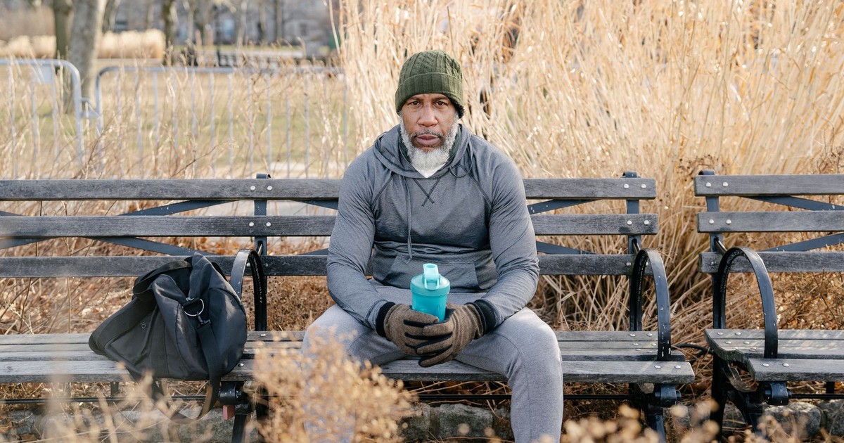 Thoughtful aged black man in warm sportswear sitting on wooden bench in park with sport bag and holding bottle of water while looking at camera in daytime