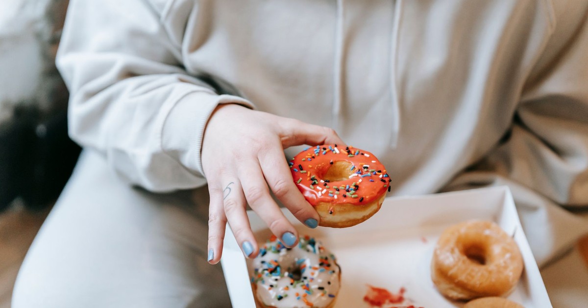 Crop unrecognizable plump female in comfy clothes picking yummy sweet donut from carton box while sitting on floor in light room
