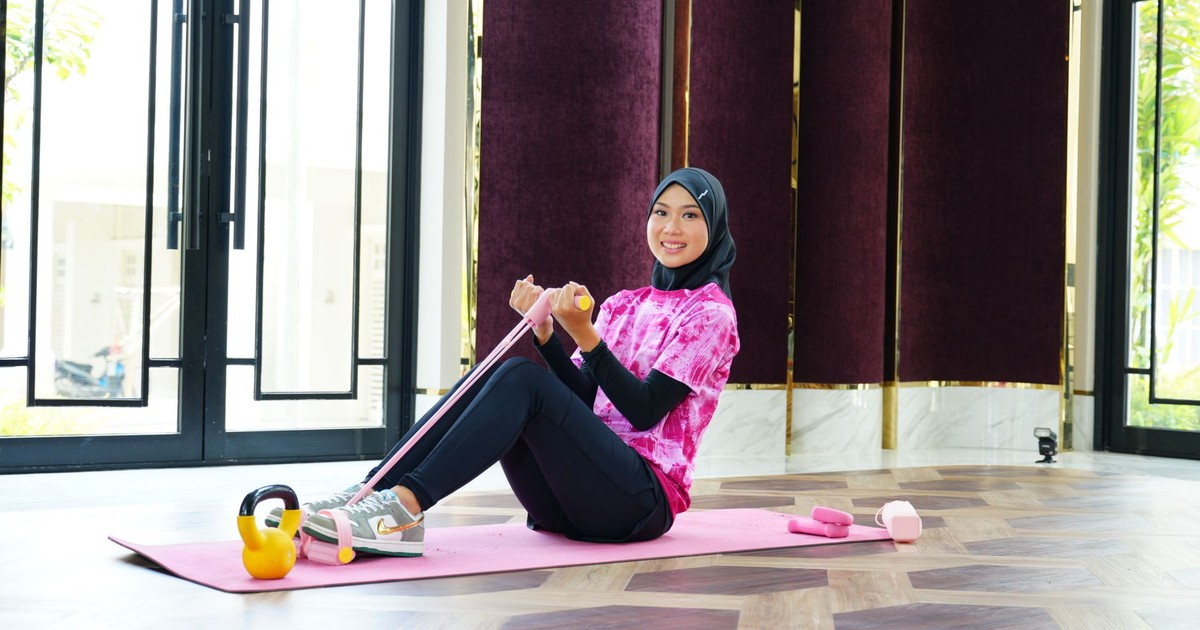 A woman in sporty attire and hijab exercises indoors using a resistance band, showcasing fitness and fashion.