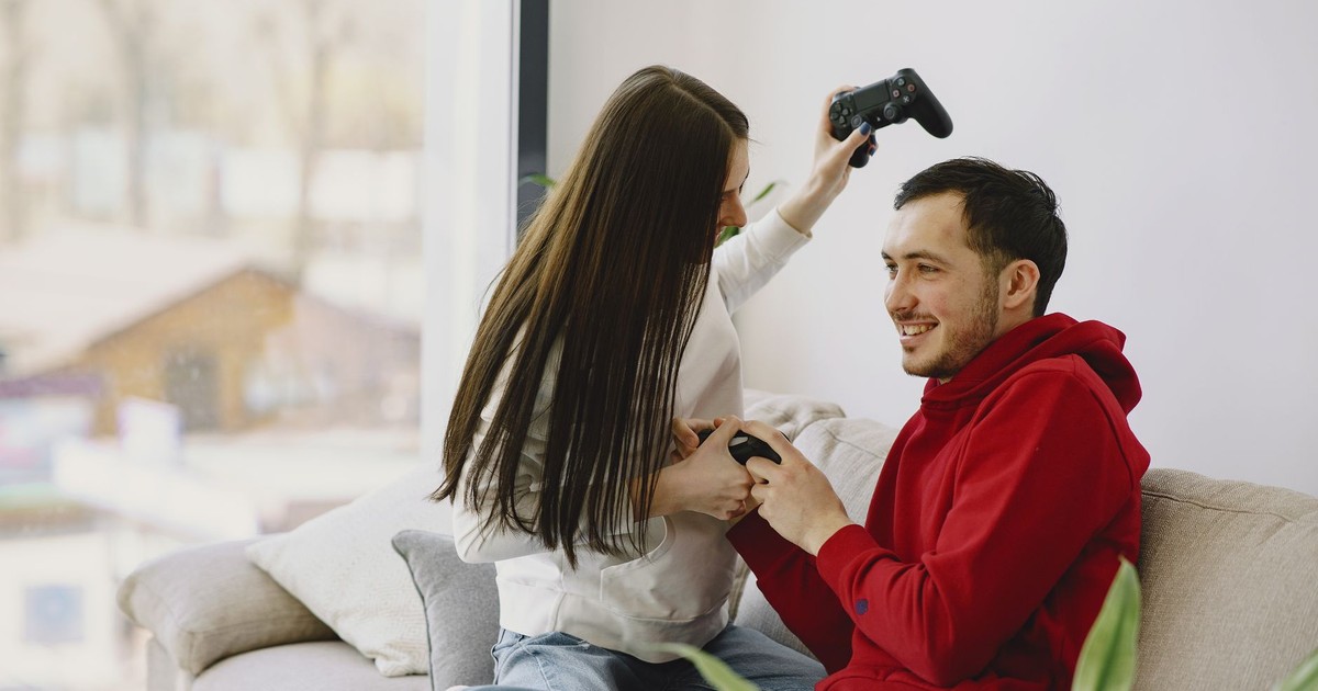 Unrecognizable woman playing with cheerful bearded boyfriend using game pad while sitting on sofa and having fun in apartment in daylight