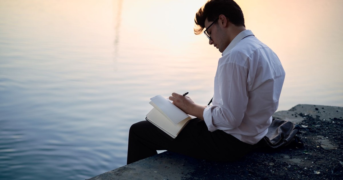 Man in white shirt writing in a notebook by the water at sunset, creating a serene and contemplative scene.