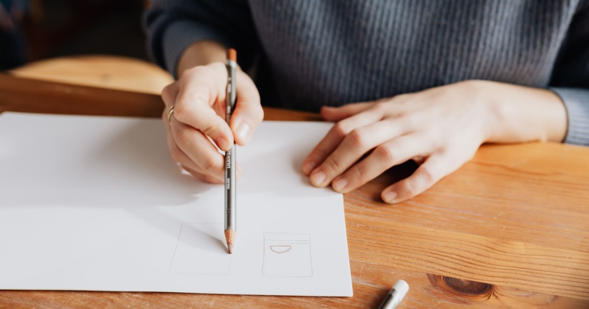 Person sketching on white paper with pencil on a wooden table, showcasing artistic creativity.