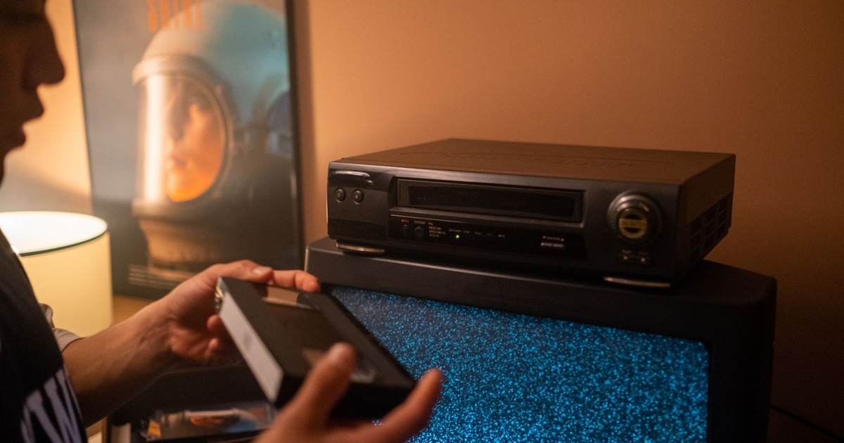 Person inserting VHS tape into video recorder with static TV in cozy room.