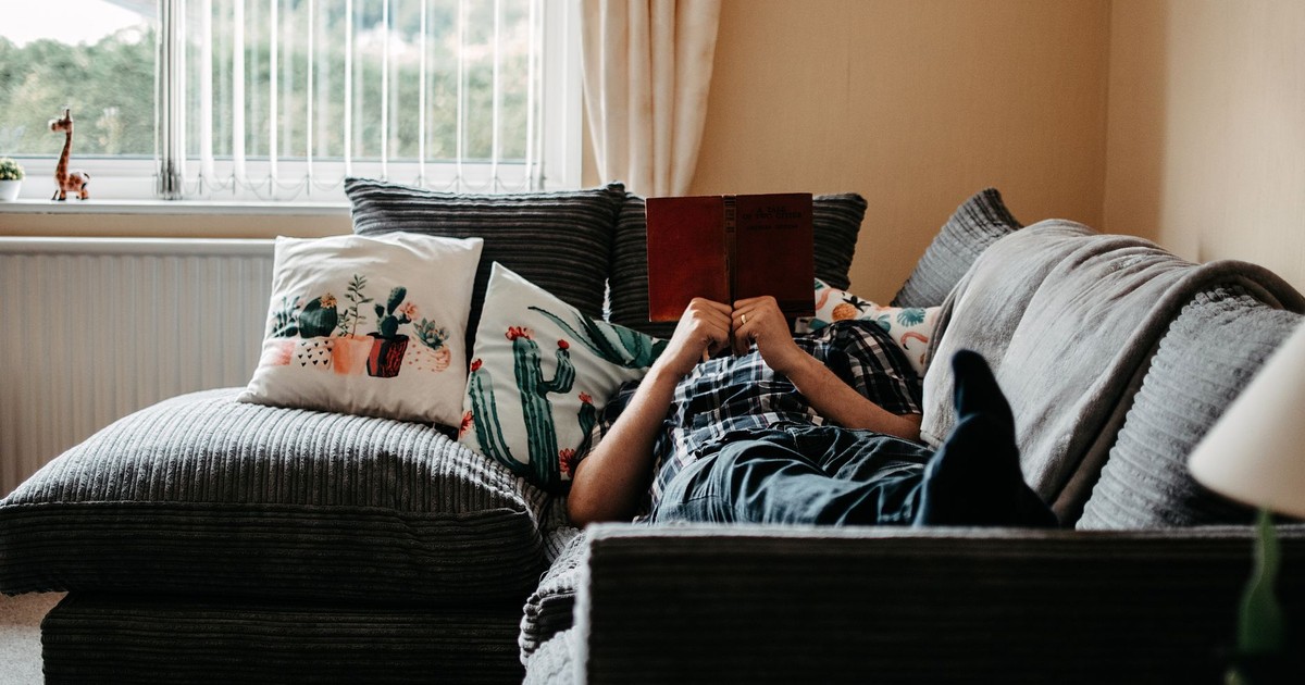 Person relaxing and reading on a cozy sofa at home, surrounded by cushions and natural light.