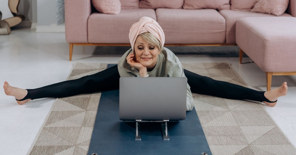 A senior woman enjoys a relaxing yoga session indoors, stretching while using her laptop.