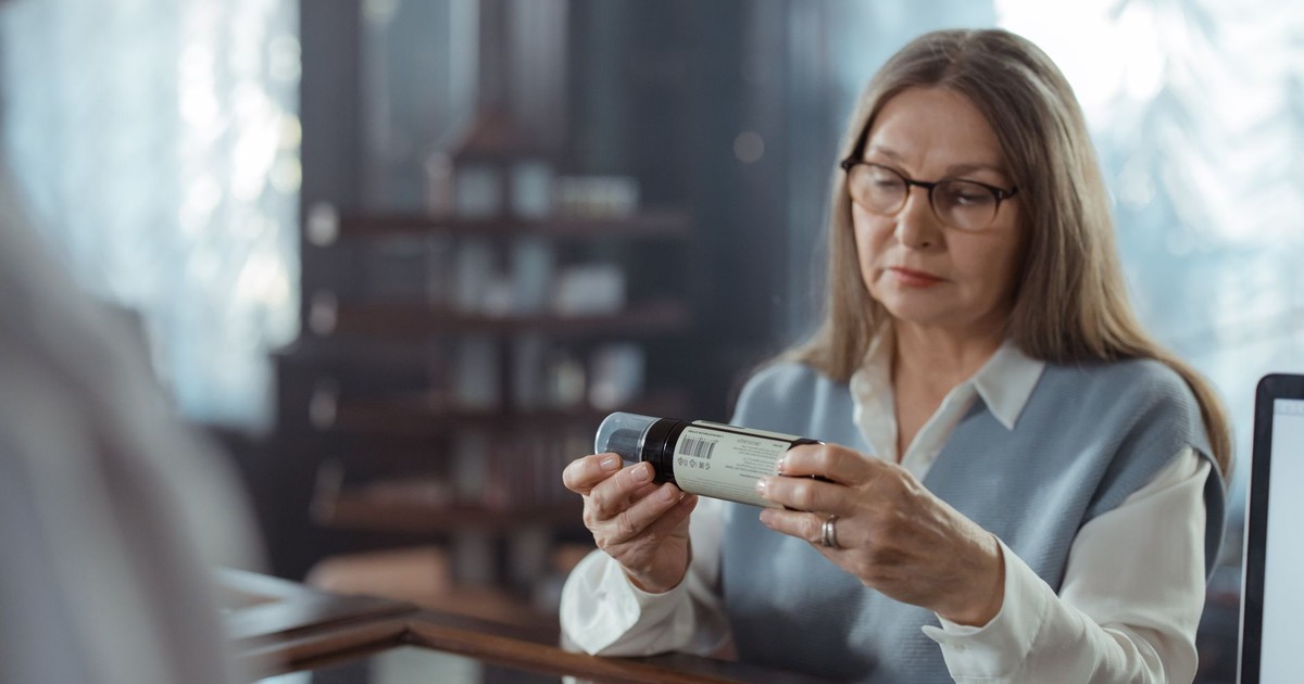 A senior woman with eyeglasses reviews a medicine bottle at a pharmacy counter indoors.