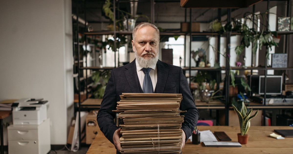 Elderly caucasian man with a beard holding piles of paperwork in a modern office environment.