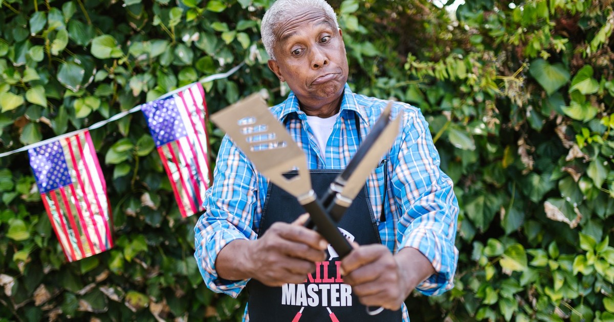 Senior man in apron holding grill tools, ready for a backyard barbecue.