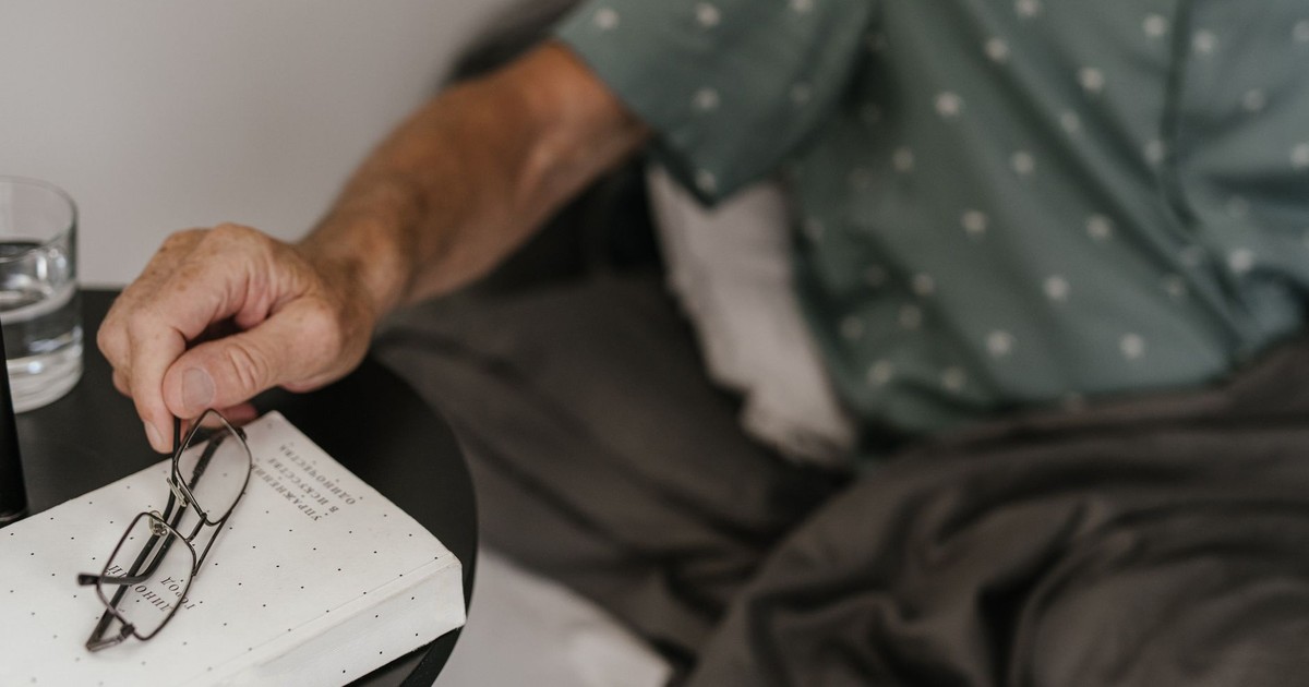 Senior adult relaxing in bed, reaching for glasses and book, creating a cozy reading moment.