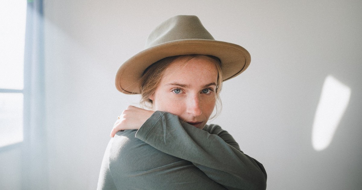 Soft portrait of a woman hugging herself in a sunlit room, wearing a stylish hat.