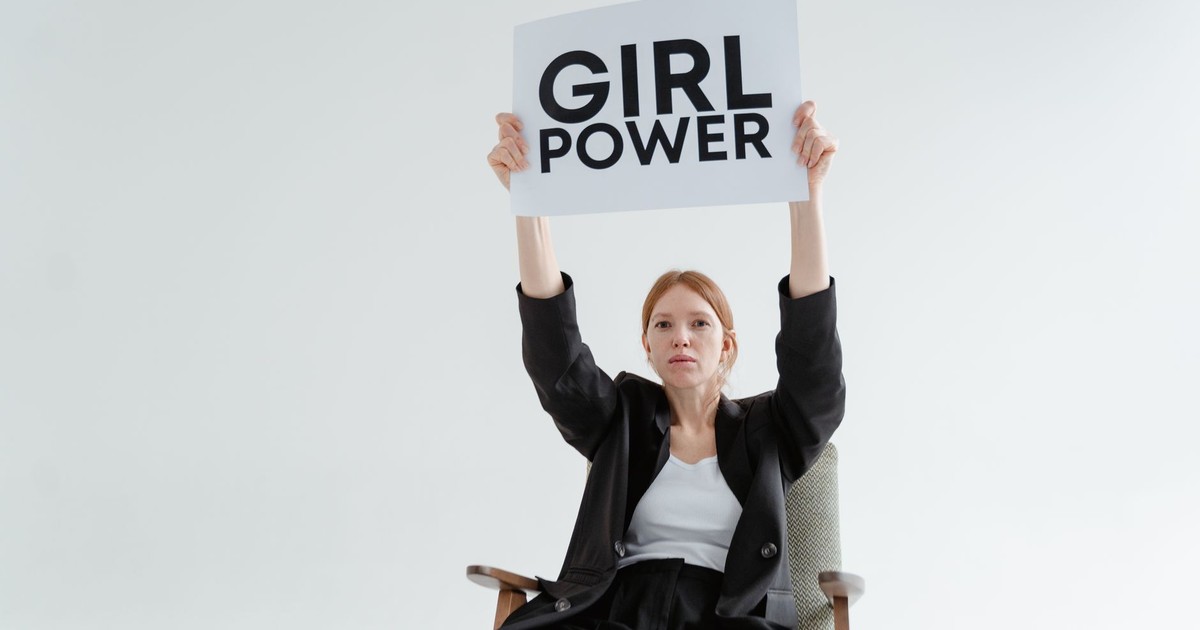 Confident woman in blazer sits with 'Girl Power' sign on white background.