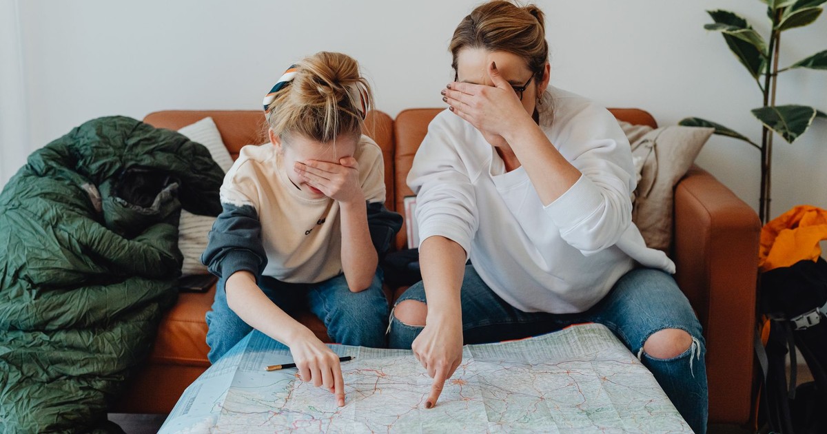 Mother and daughter covering eyes while pointing at map on couch, planning a trip.