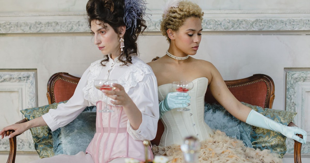 Two stylish women in Victorian attire enjoy wine in an elegant setting.