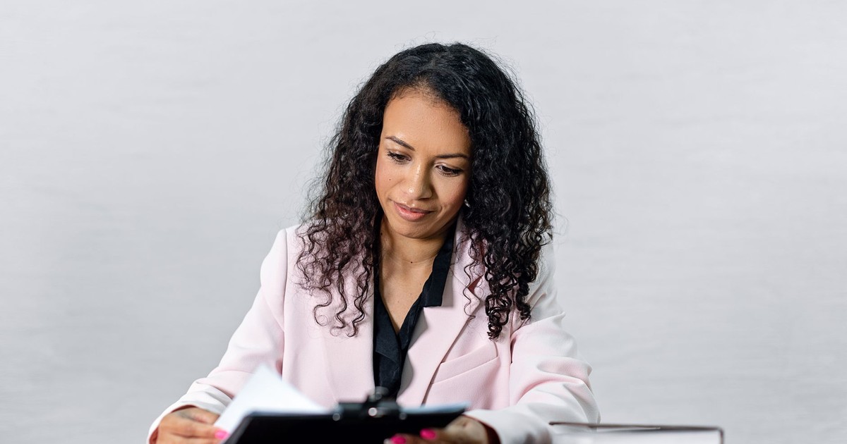 Confident woman with curly hair in a formal attire reviewing documents at a desk.