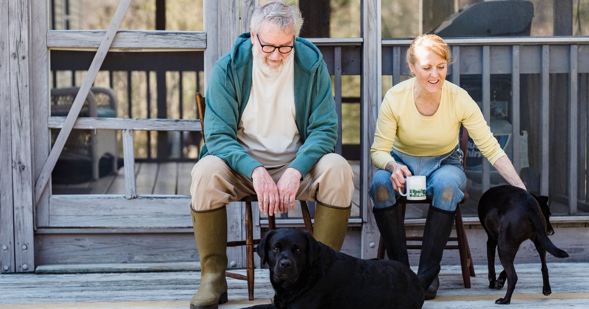An elderly couple sits with their two dogs on a wooden porch, sharing a peaceful moment outdoors.