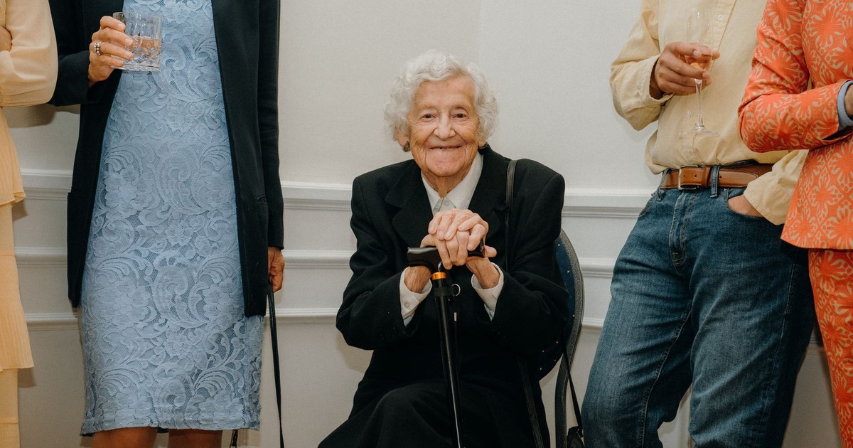 Senior woman smiling at a formal gathering, surrounded by elegantly dressed people.